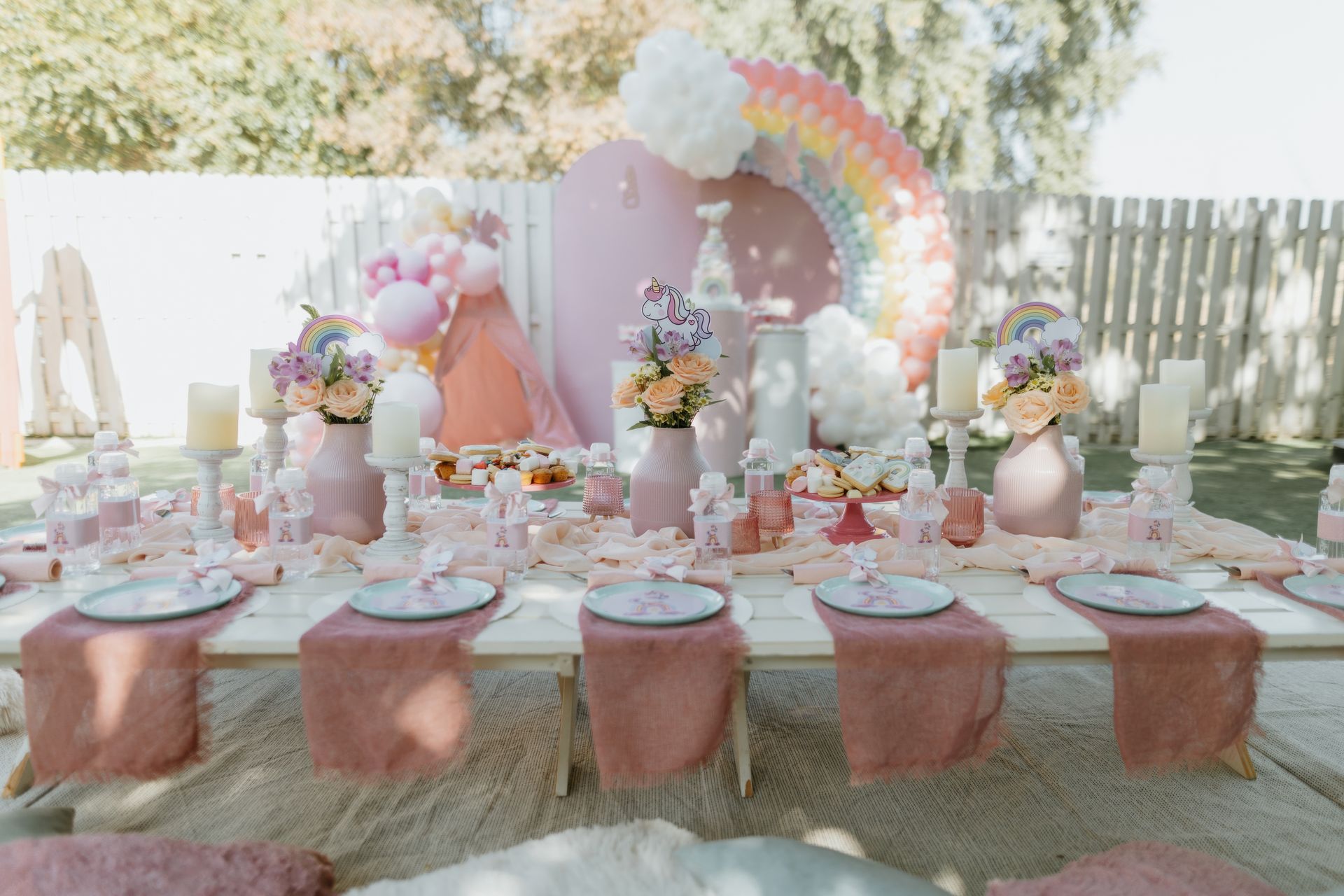 Pastel pink dessert table with plates, balloons, flowers, and a rainbow backdrop in a garden setting