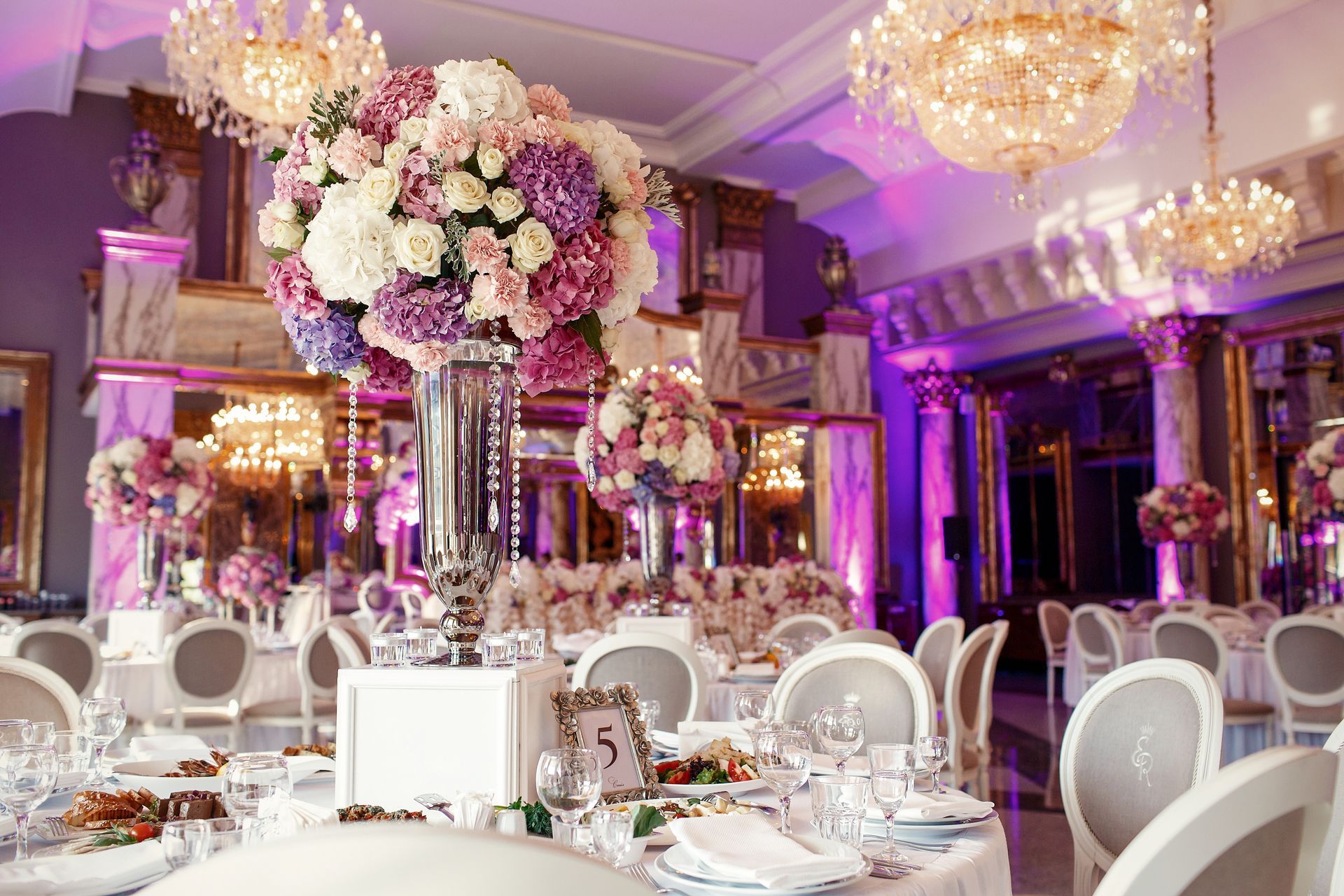 Elegant banquet hall with white-and-gold table settings, chandeliers, and tall pink-and-white floral centerpieces