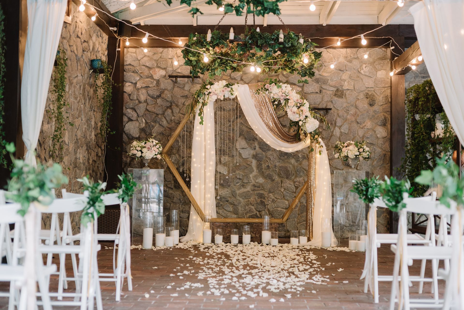 Decorated outdoor wedding altar with white chairs, floral drapes, and stone wall backdrop