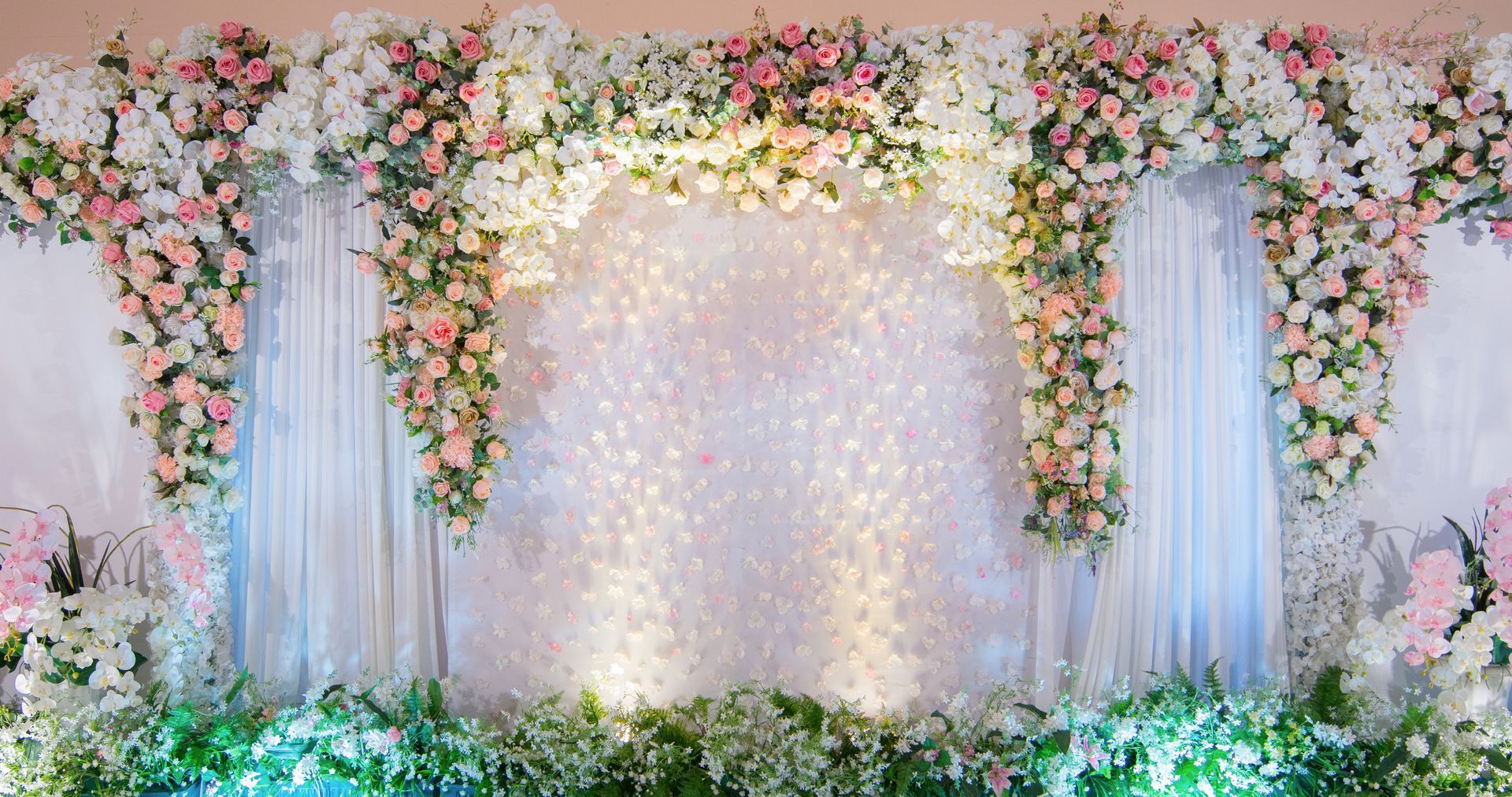 Pastel floral wedding backdrop with white drapes, twinkling lights, and greenery.