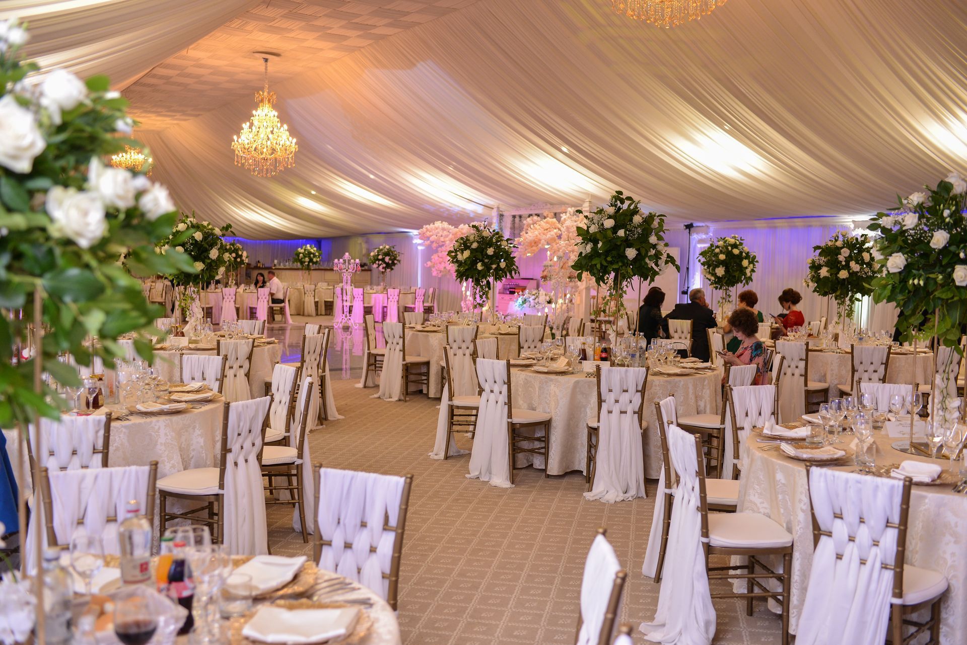 Elegant banquet hall with round tables, white chairs, floral decor, and warm chandelier lighting under a draped tent ceiling