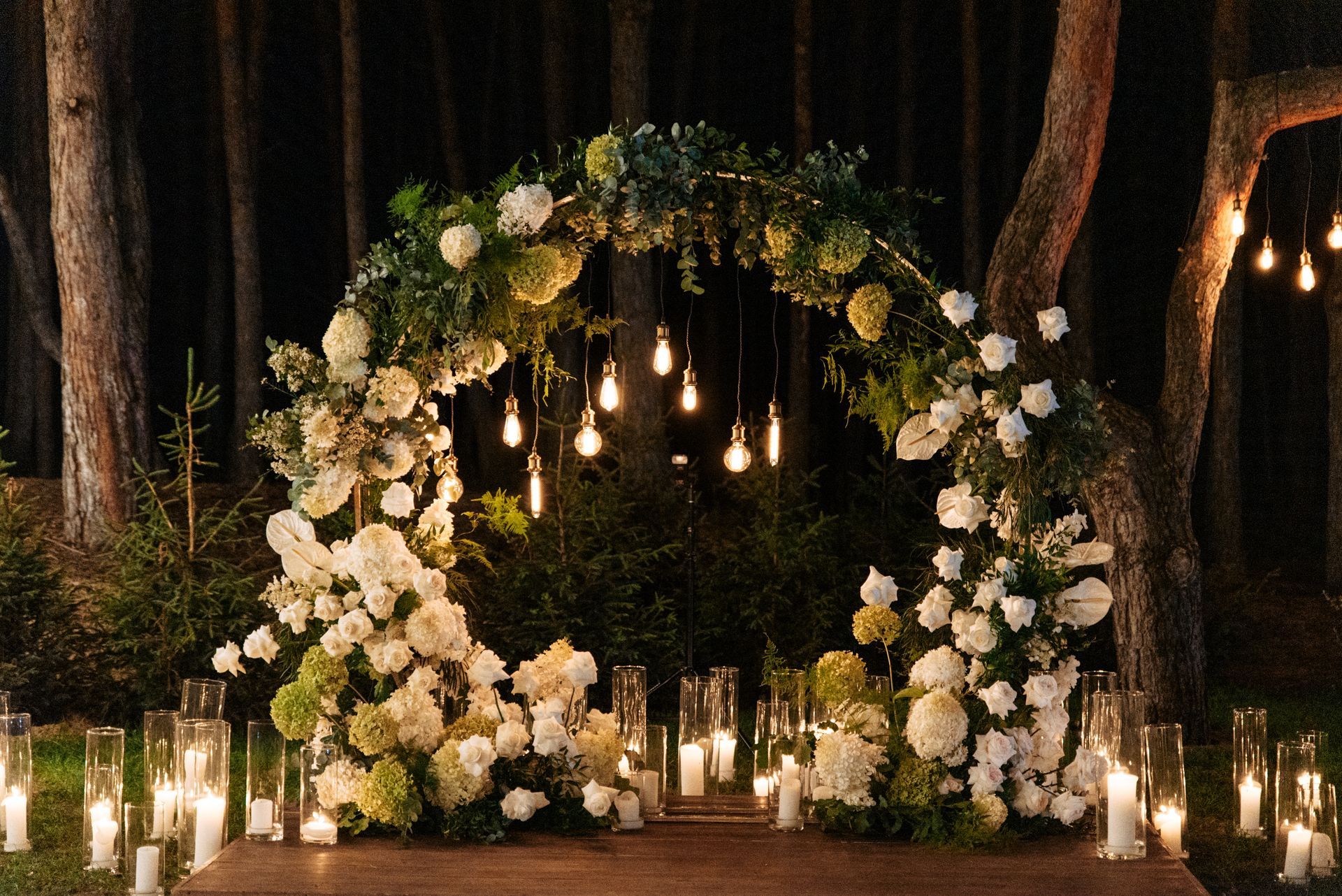Floral wedding arch with white flowers and hanging lights in a dark wooded setting