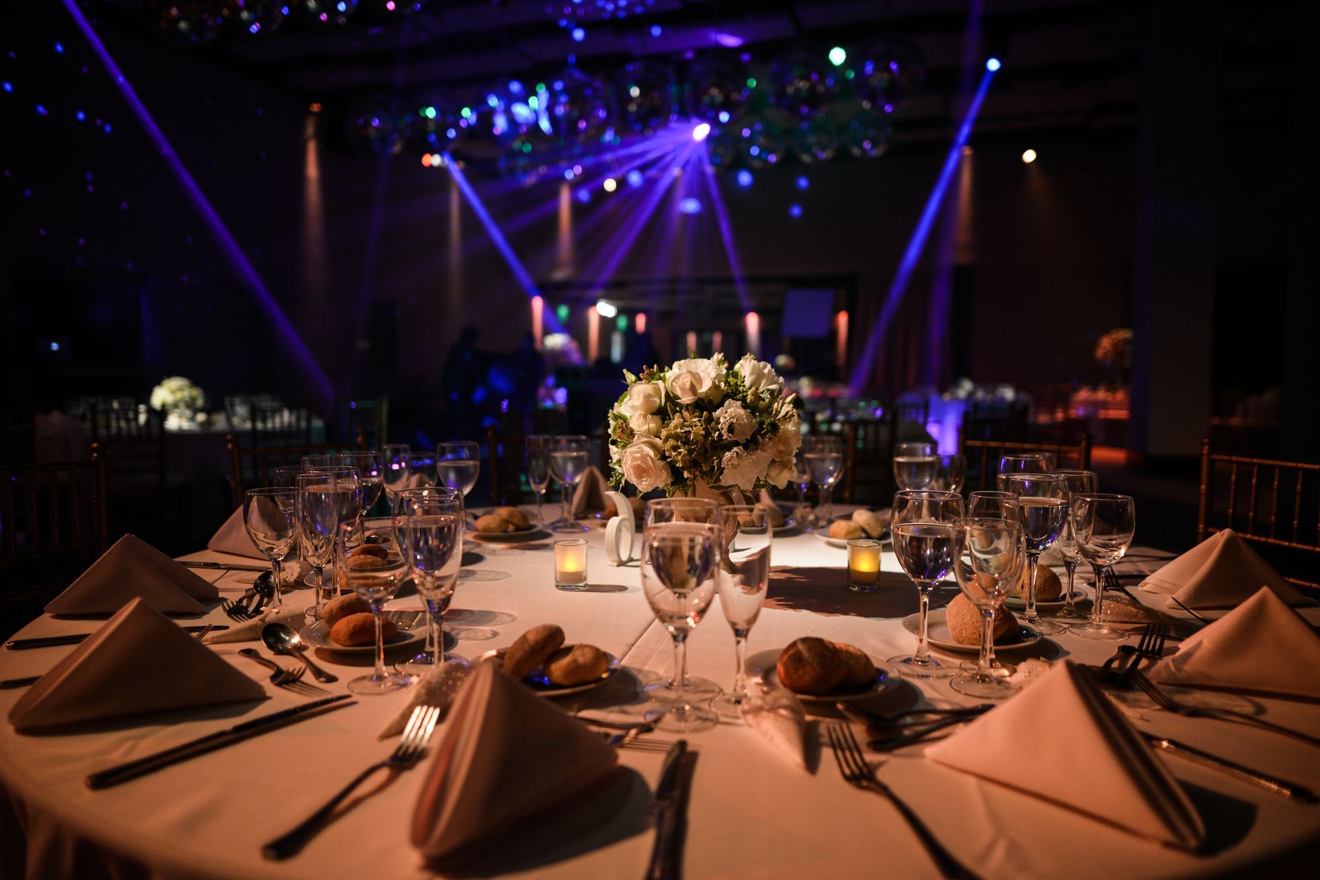 Elegant banquet table with floral centerpiece and folded napkins in a dimly lit event hall with purple stage lights