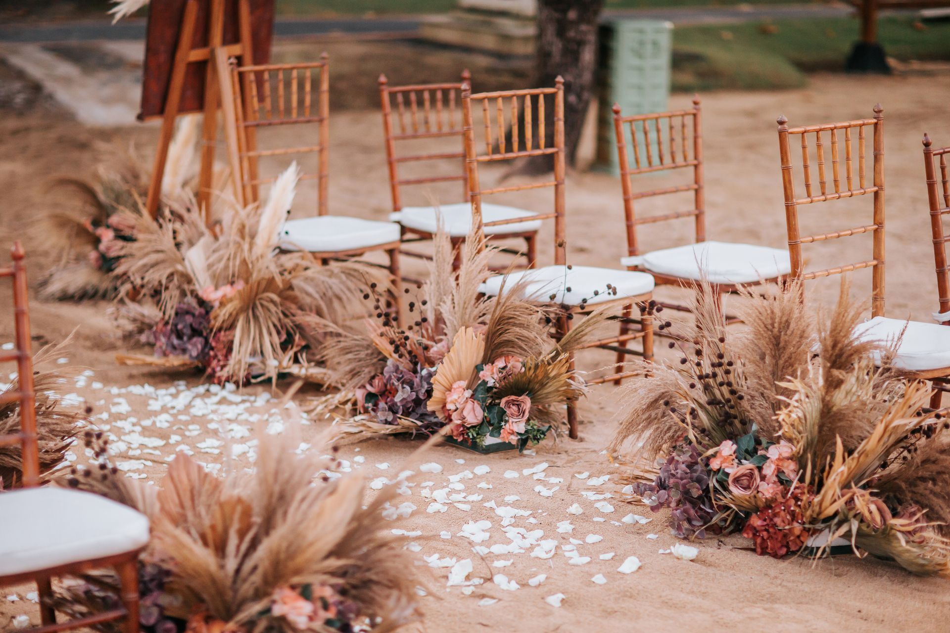 Boho outdoor ceremony aisle with gold chairs, pampas grass, dried florals, and scattered white petals