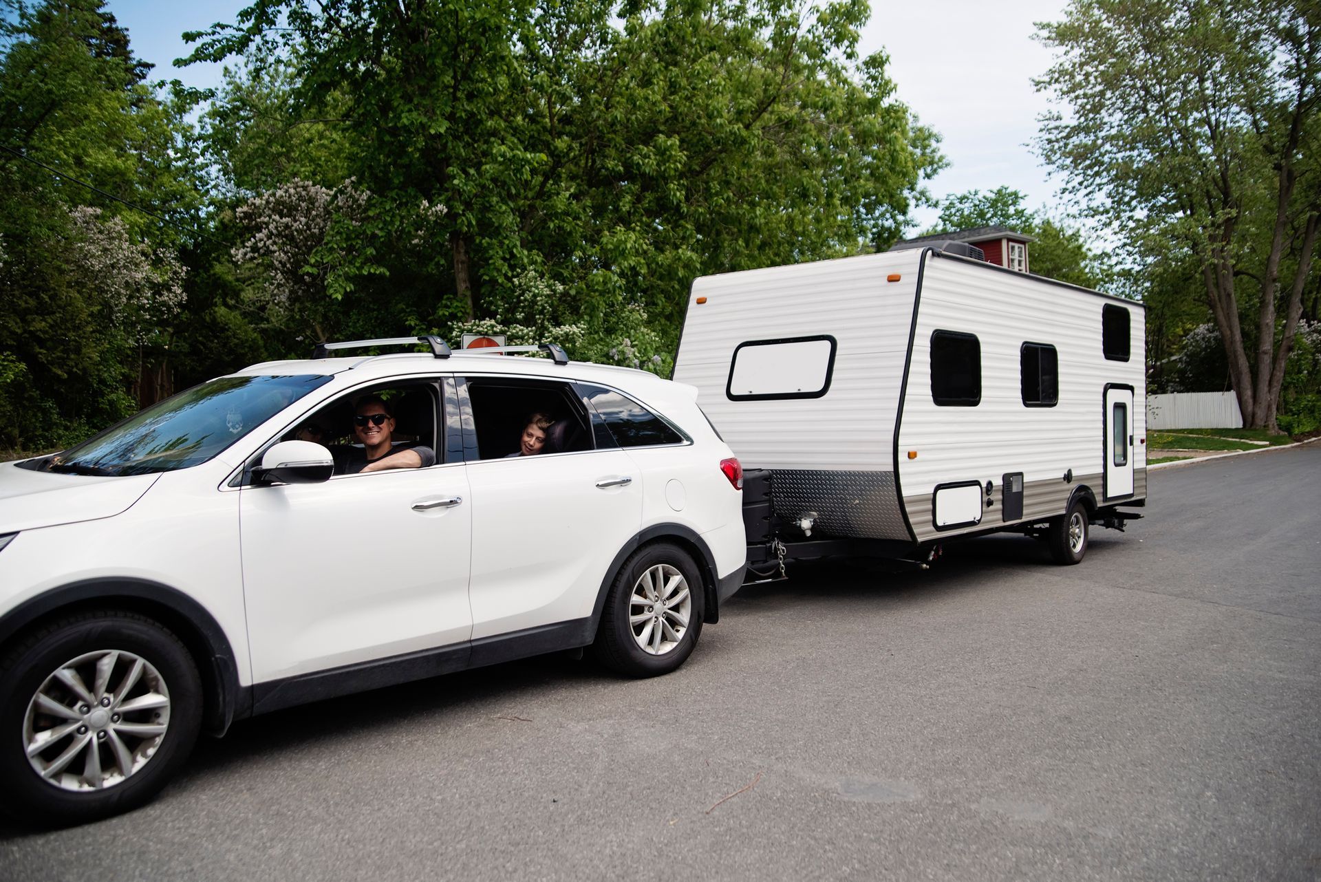 White SUV towing a travel trailer on a tree-lined road.