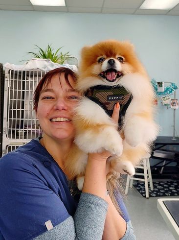 Woman holding a fluffy orange and white Pomeranian dog in a grooming shop, both smiling.
