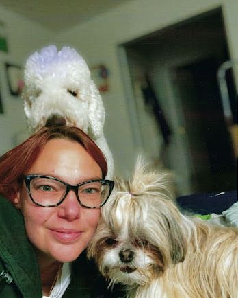 Woman grooming a white dog at a grooming table, using clippers. Mirror reflects the groomer and room.
