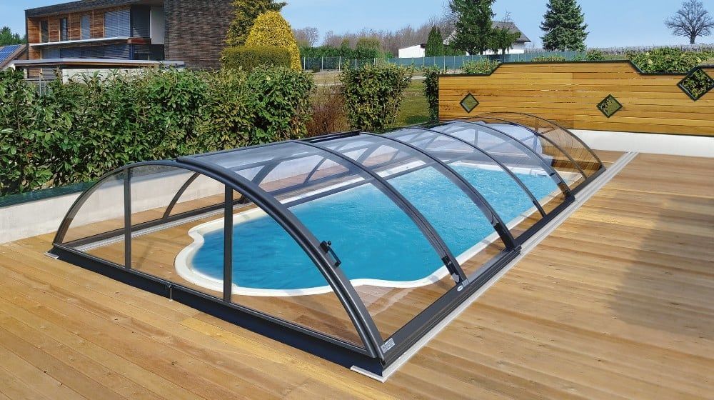Überdachter Swimmingpool im Garten mit geschwungener, transparenter Umrandung auf einer Holzterrasse, neben einem Haus und einer Hecke
