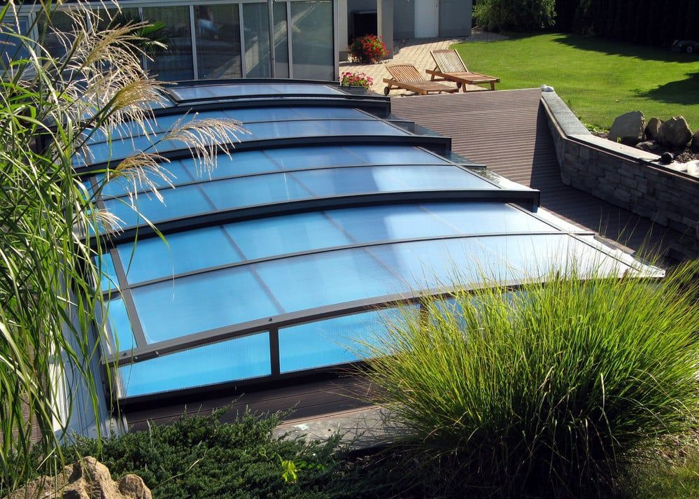 Überdachter, blau getönter Glaspool neben einer angelegten Terrasse und einem Rasen