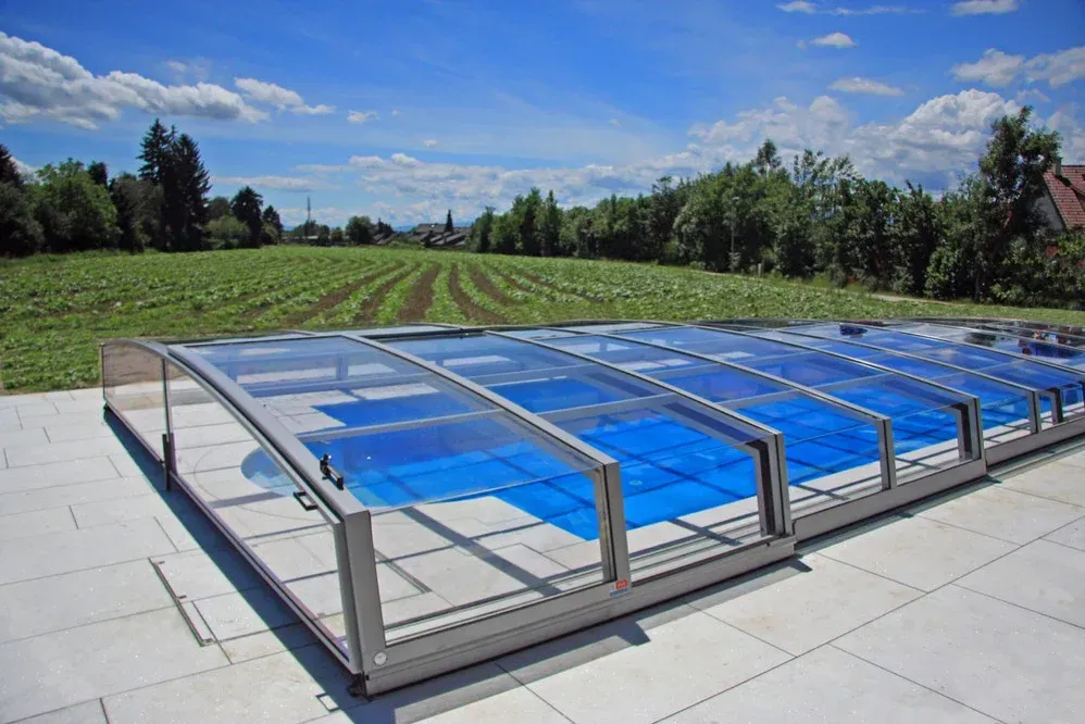Ein rechteckiger, verglaster Pool oder Whirlpool neben einer gefliesten Terrasse, im Hintergrund Felder und Bäume.