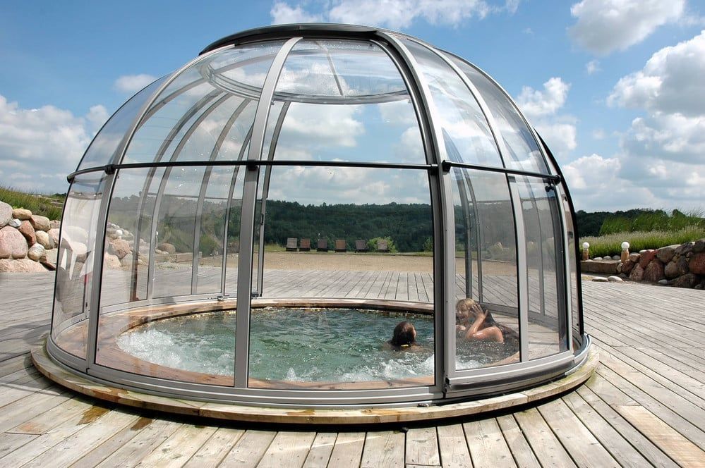 Ein verglaster Außenpool auf einer Holzterrasse, in dem eine Person unter einem bewölkten Himmel schwimmt