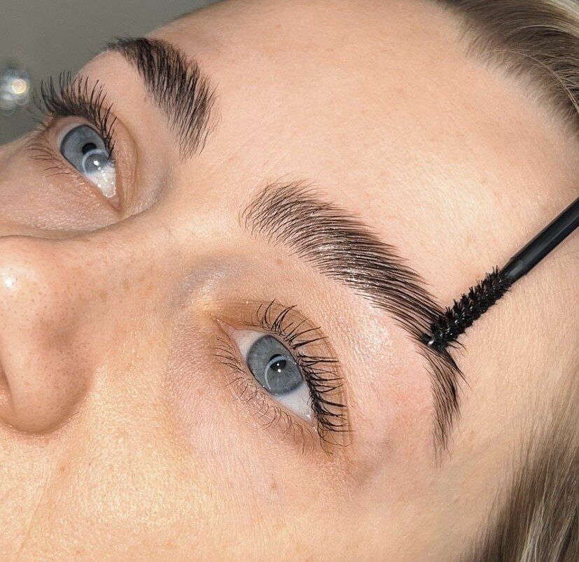 Close-up of a woman with blue eyes, getting her eyebrows brushed and styled, likely a brow lamination.