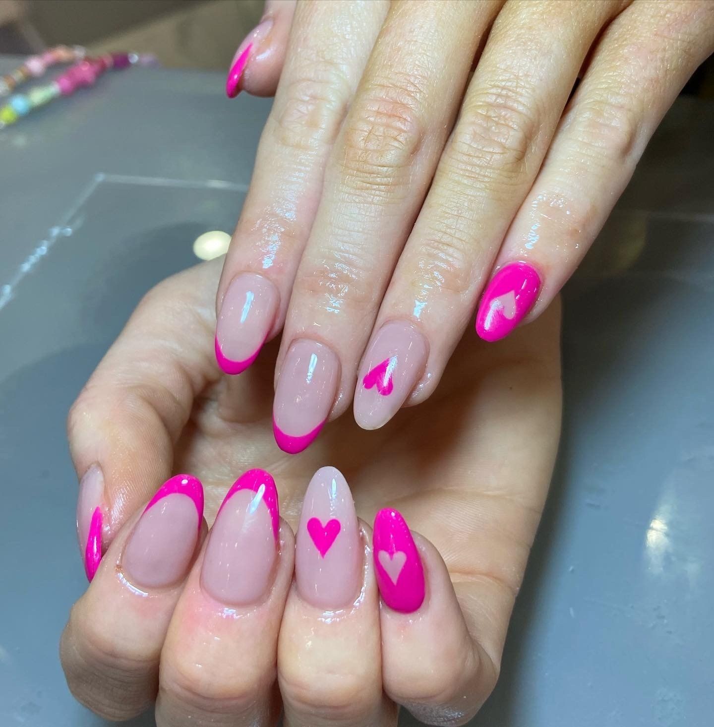 Hands with pink heart-themed nail art: French tips and heart accents on a light pink background.