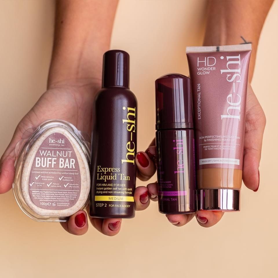 Hands holding four He-Shi self-tanning products: Walnut Buff Bar, Liquid Tan, self-tanning mist, and HD Wonder Glow.