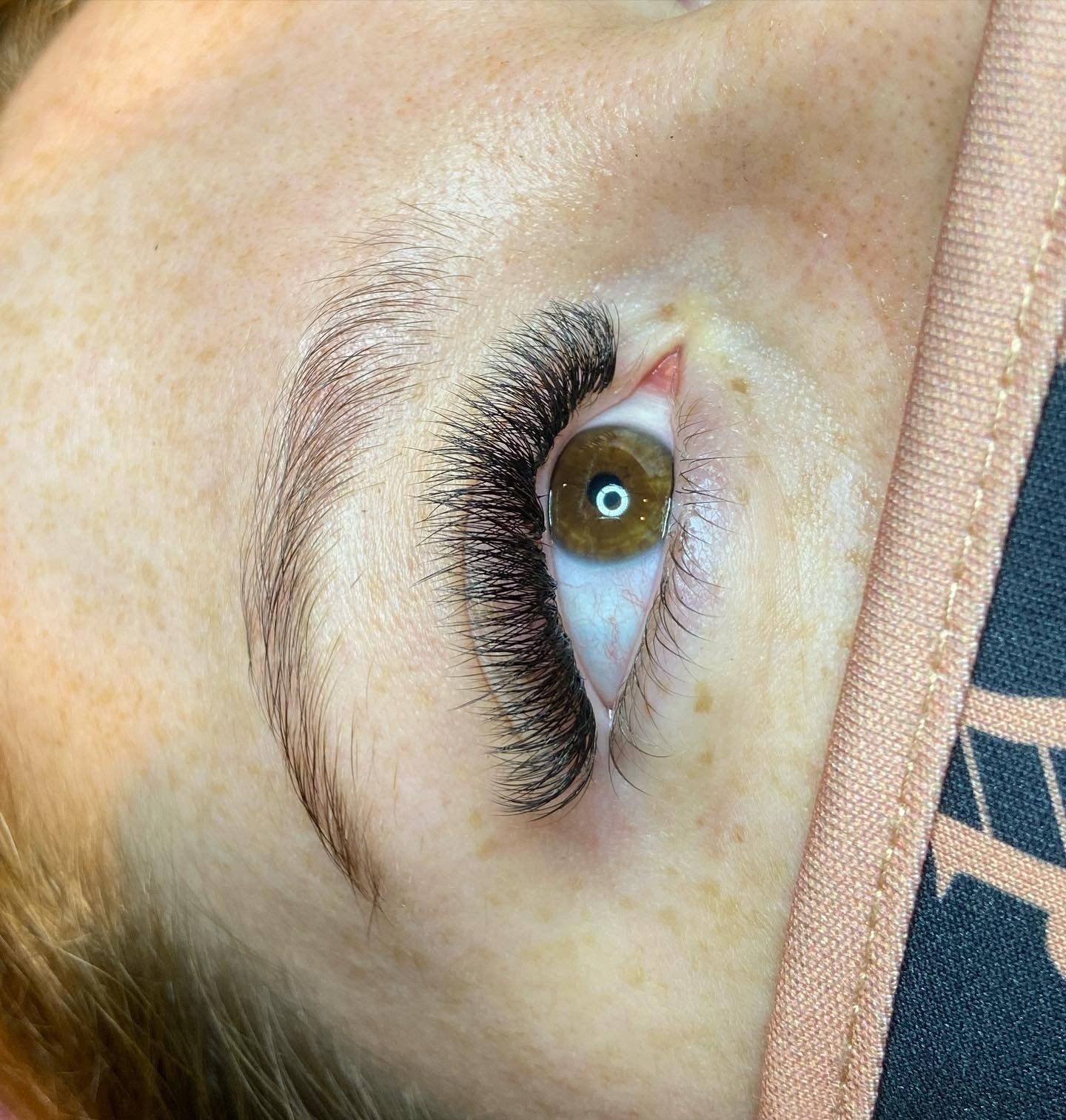 Close-up of an eye with thick, black eyelash extensions, light brown eyebrows, and a brown iris.