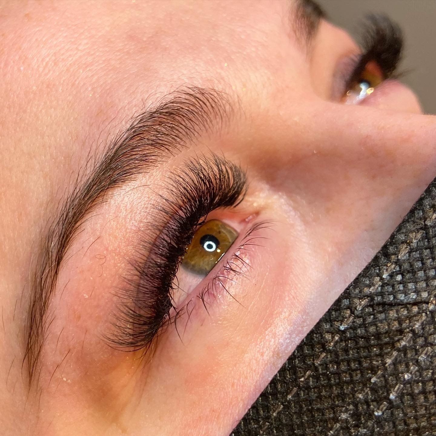 Close-up of a person's eye with thick, dark eyelashes and well-groomed eyebrow. The eye is brown, skin is light.