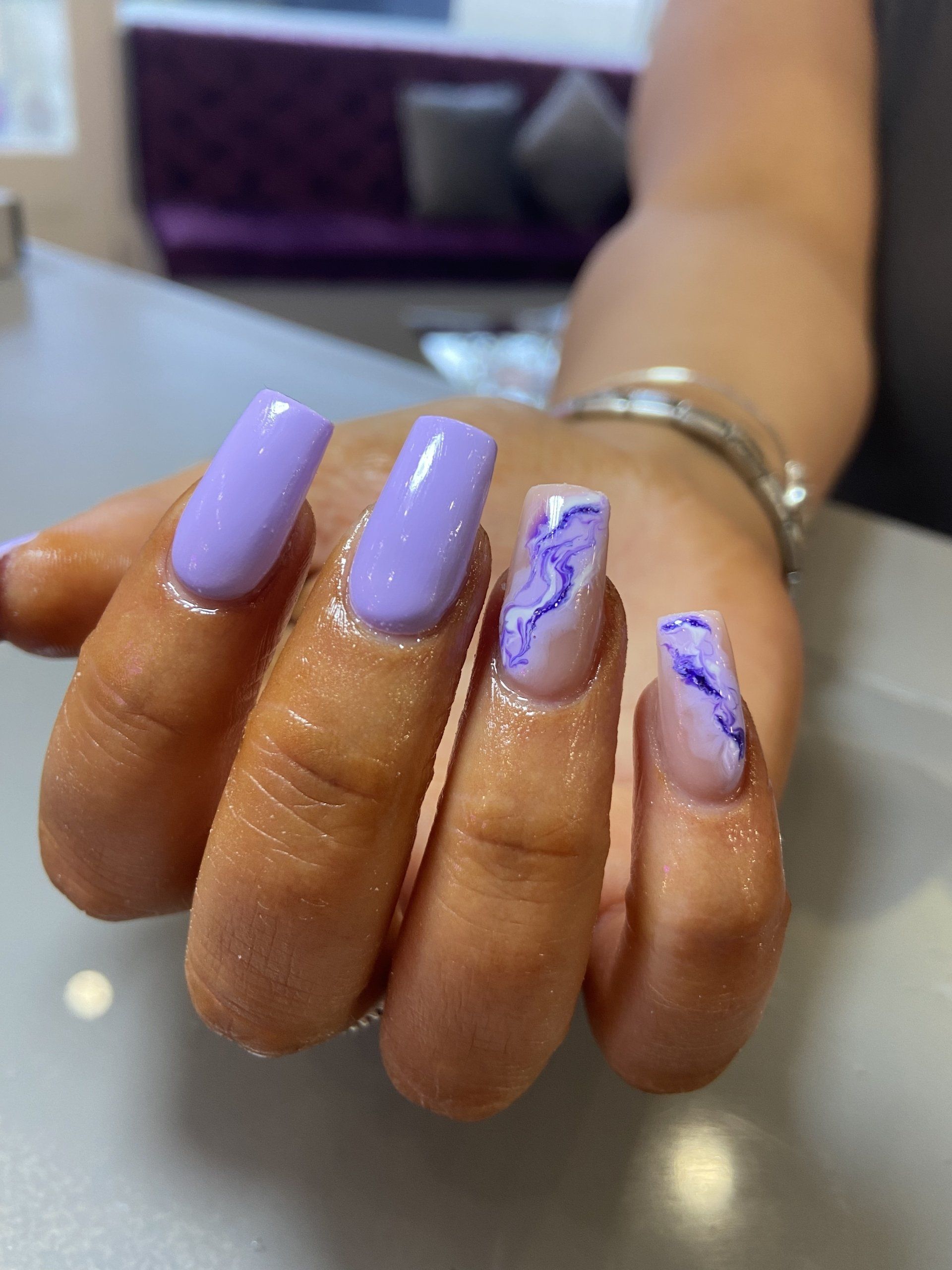 Hand with light purple square-shaped nails, one with a marble design, against a neutral background.