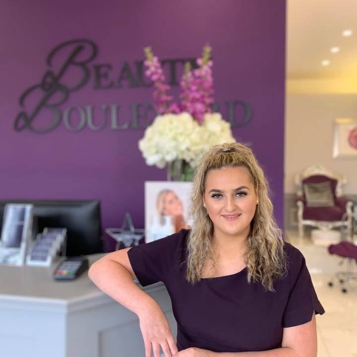 Woman with blonde hair leans on a salon reception desk in front of a purple wall with a business sign.