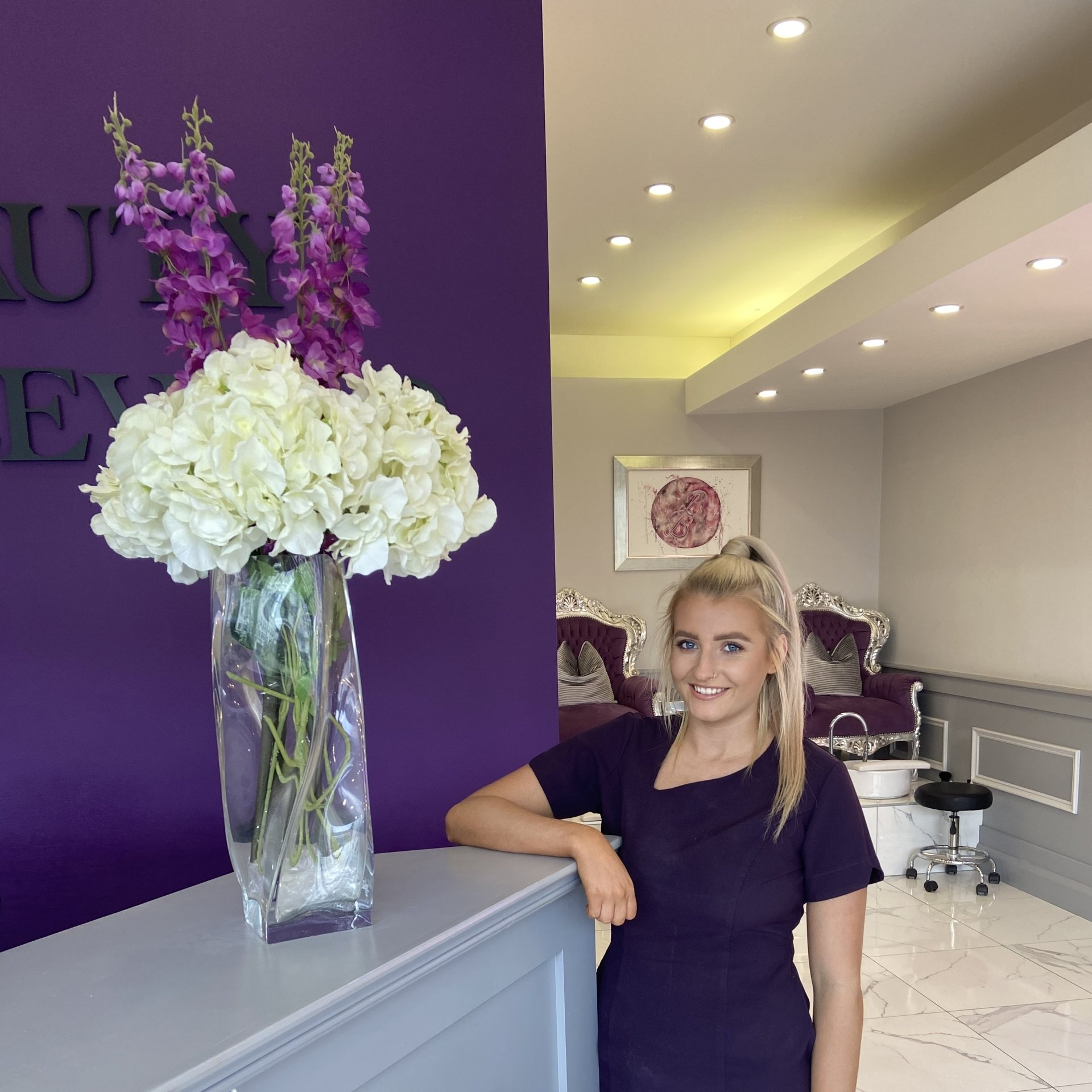 Woman in salon, smiles, leans on a counter with flower arrangement. Purple wall, silver decor.