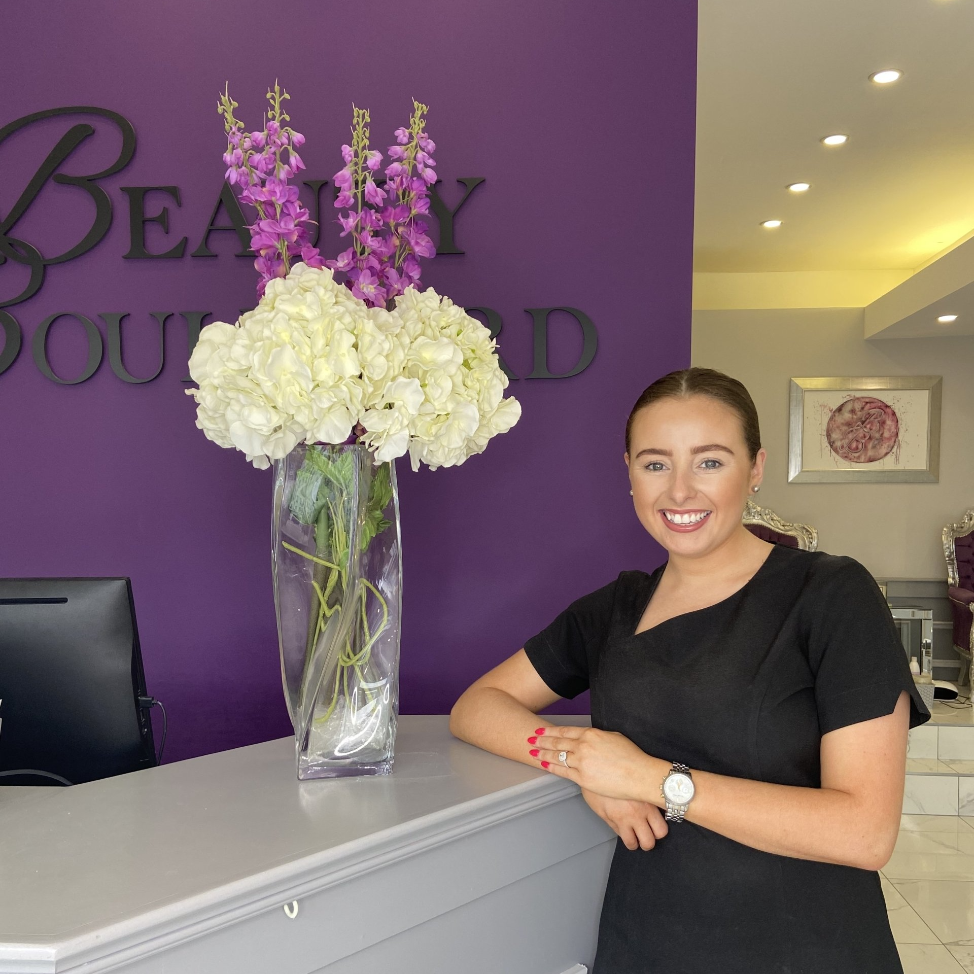 Woman at desk smiling in front of a purple wall with flowers.