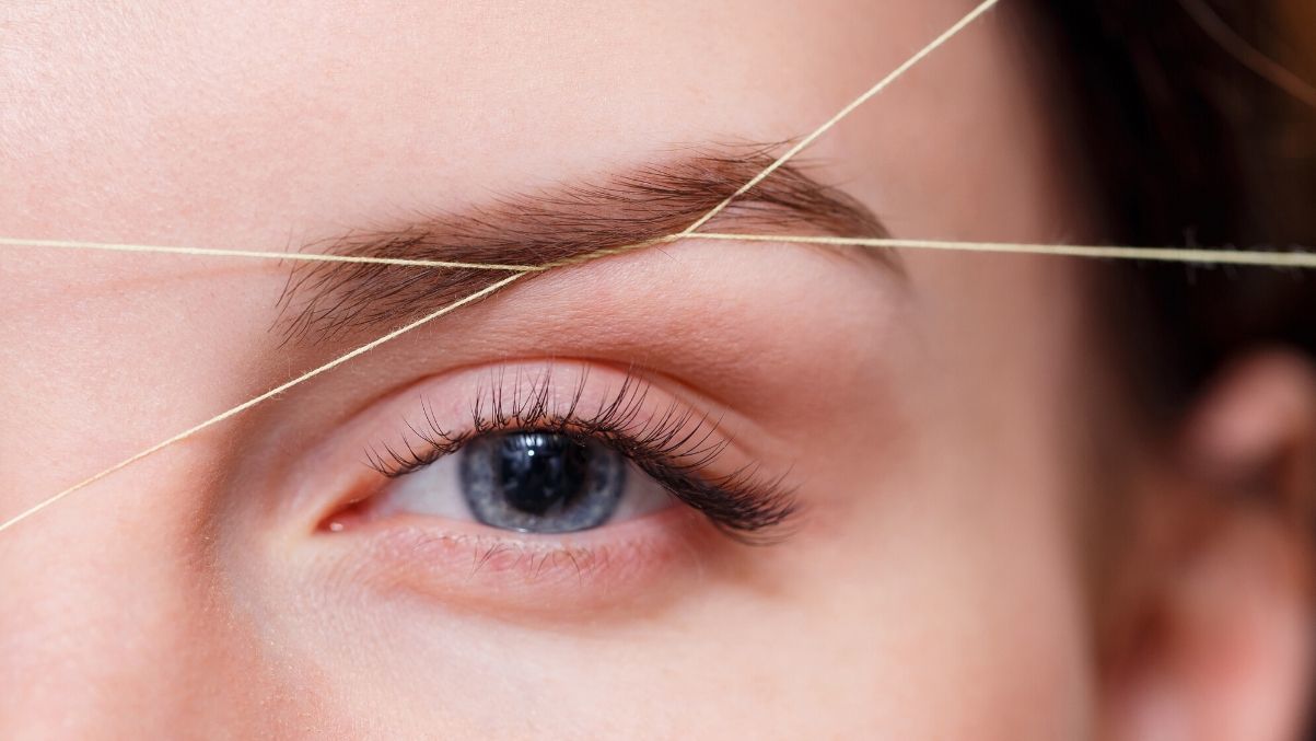 Woman's eye and eyebrow being shaped with threading. Light skin, blue eye, brow is brown.
