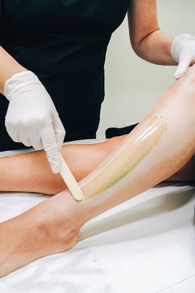 Person applying wax to leg with a spatula, wearing gloves in a salon.
