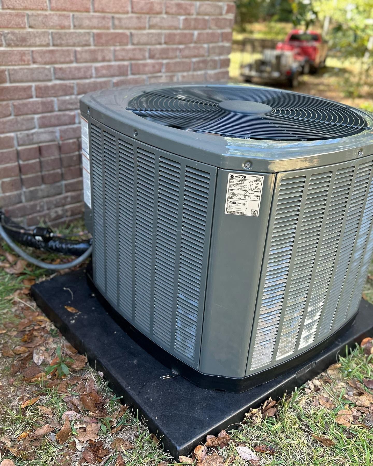 Outdoor central air conditioning unit beside a brick wall on a black pad.