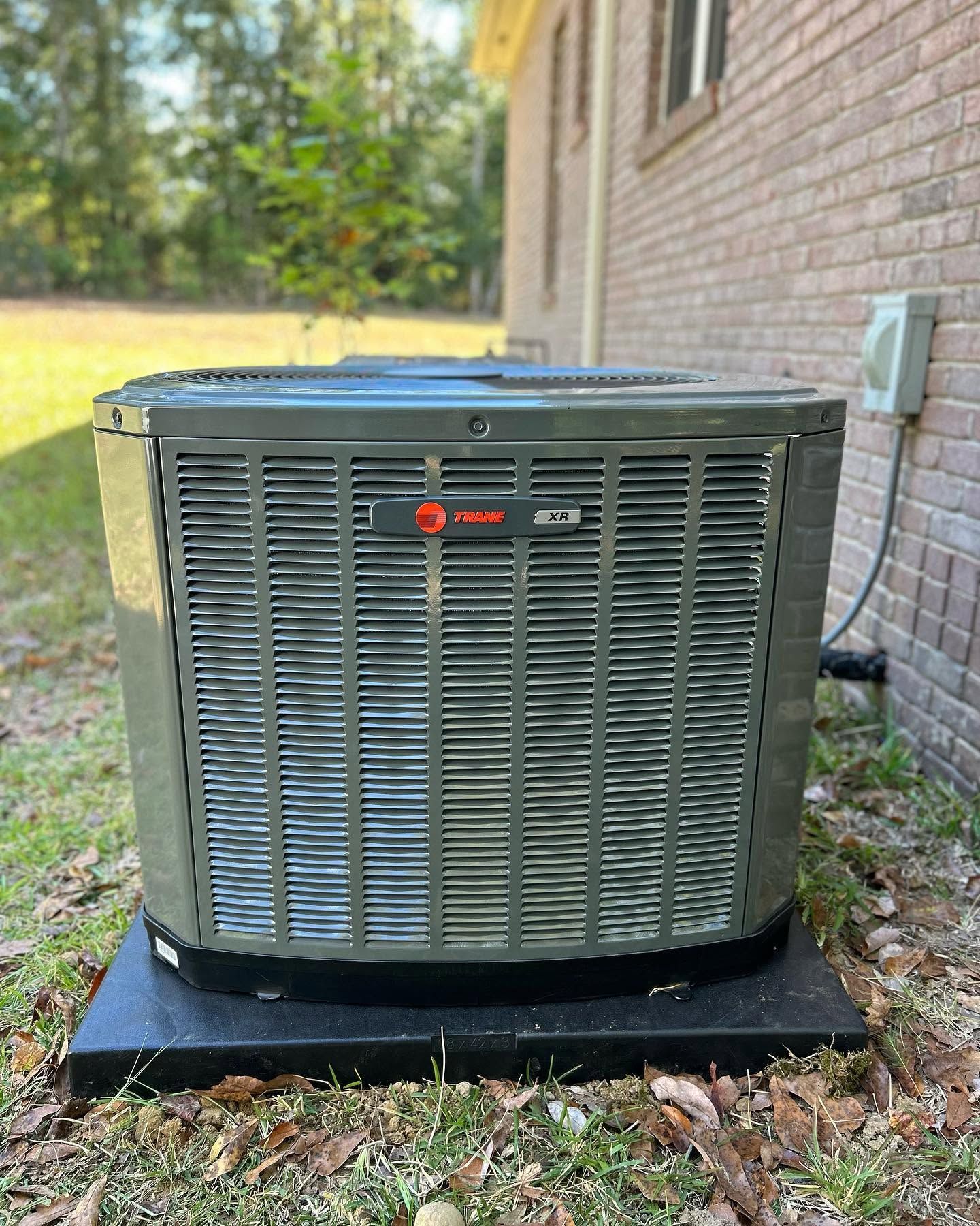 Outdoor HVAC condenser unit beside a brick house on a black pad with leaves around it