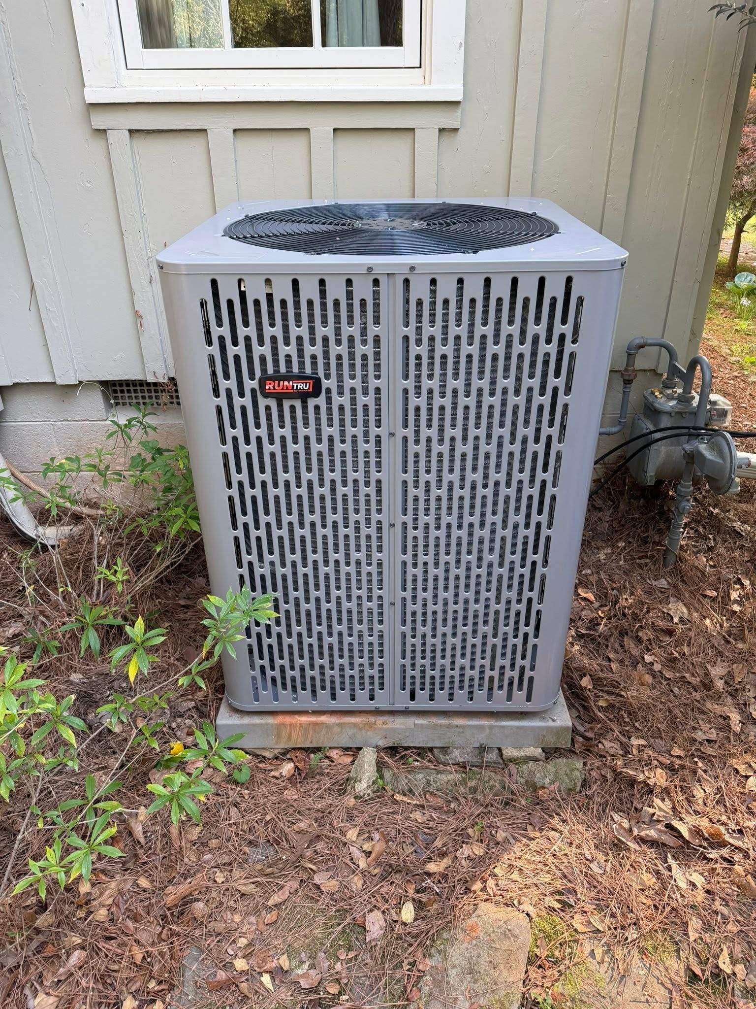 Outdoor air conditioner unit beside a house wall, surrounded by mulch and weeds