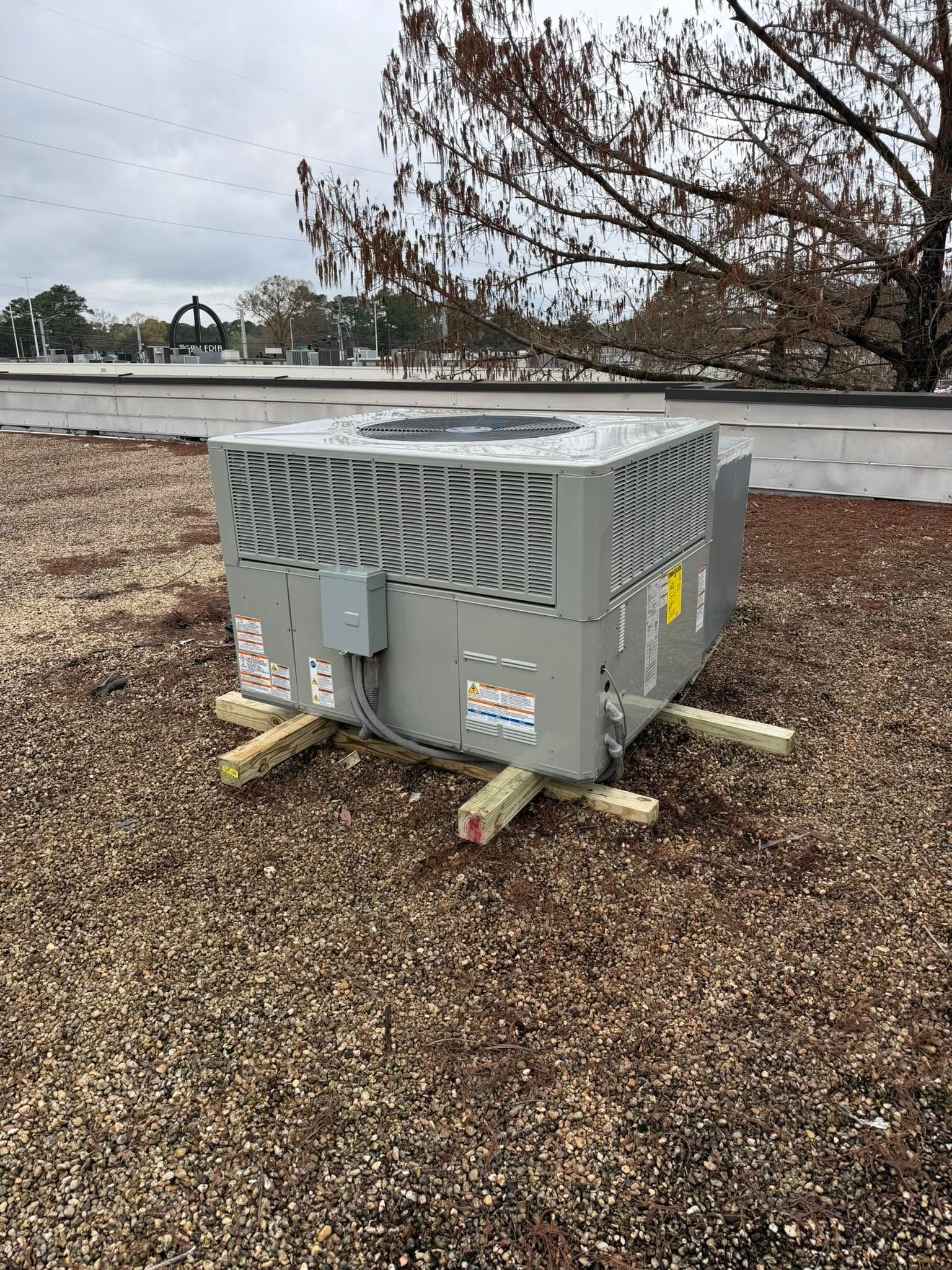 Gray rooftop HVAC unit on gravel, with leafless tree branches in the background.