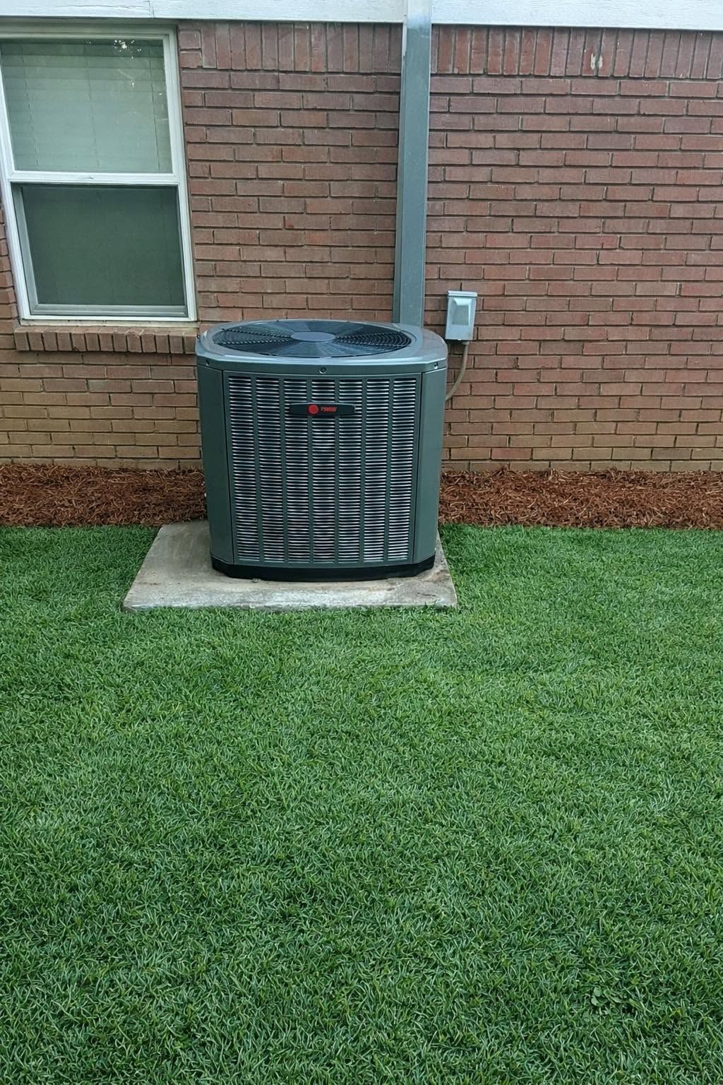 Outdoor HVAC unit beside a brick house wall and window, on a concrete pad over green grass.