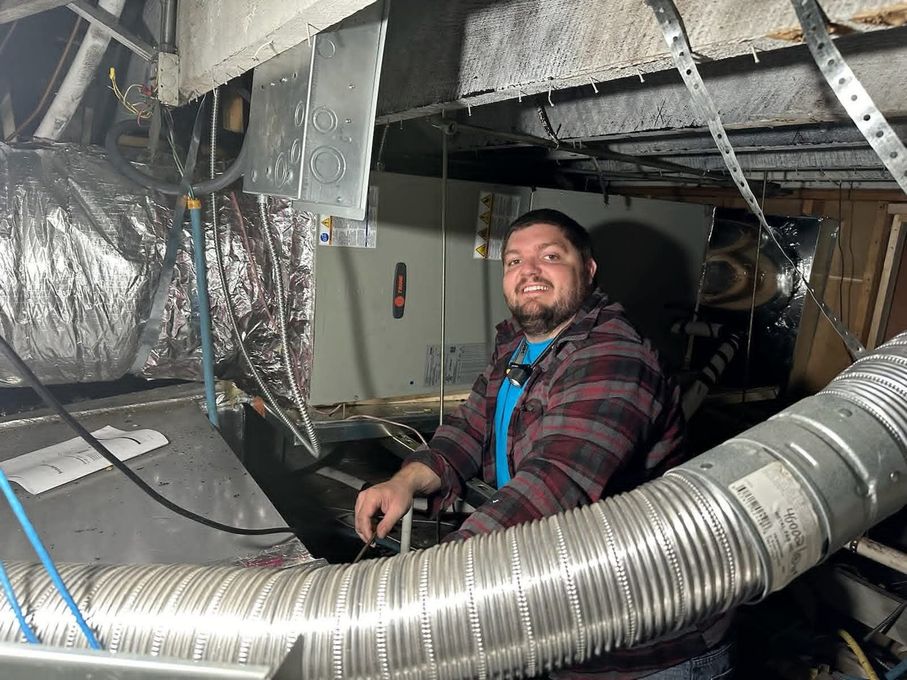 Worker smiling in a crawl space among ducting and cables, wearing a plaid shirt and blue harness.