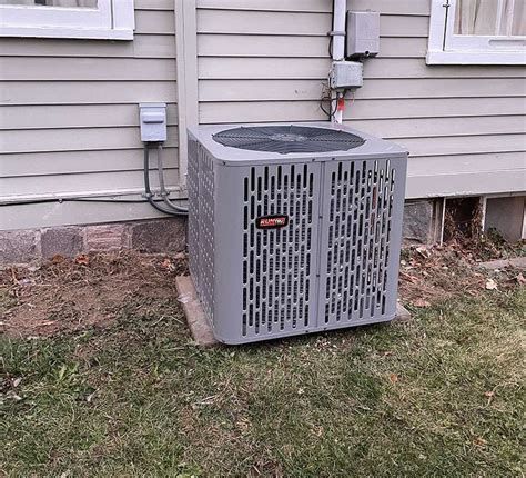 Outdoor AC unit beside a house wall, with gray siding and a grass yard.