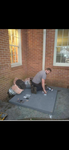 Two people kneeling on a gray mat outside a brick building, working on a roof or platform.