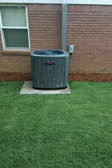 Outdoor air conditioner unit beside a brick house wall and window on a concrete pad, with green grass