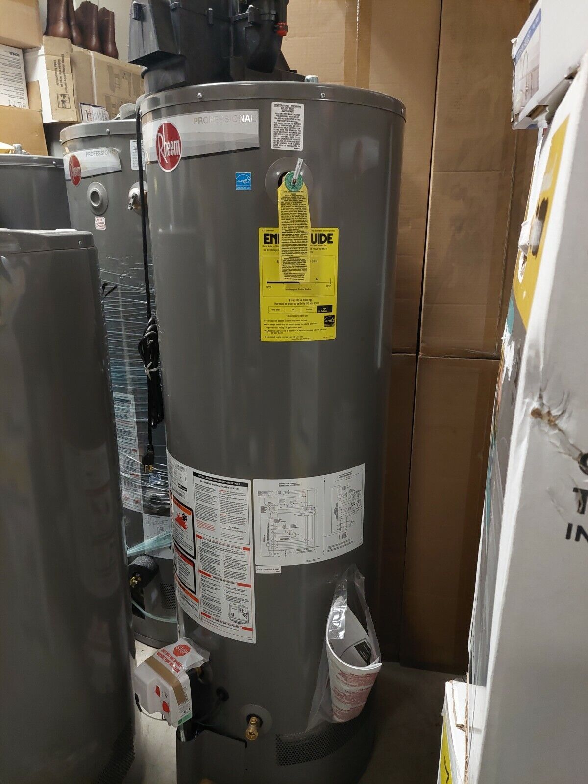 Gray water heater in a storage area, with warning labels and boxes stacked around it.
