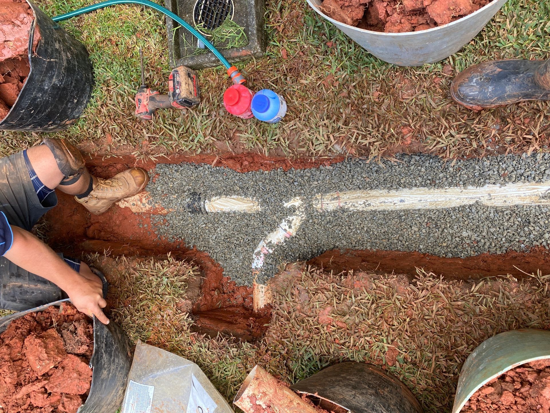 Hands Digging in Red Soil Around a Marked Trench with Buckets and A Hose Nearby — Port Macquarie Plumbing In Port Macquarie, NSW