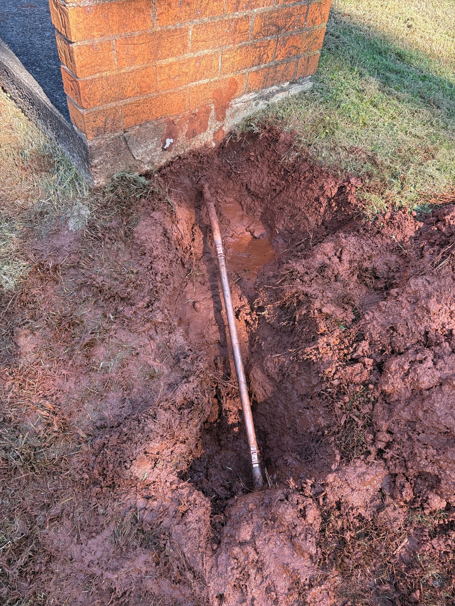 Brick Wall with A Trench in Dirt and A Metal Pipe or Cable Running Into the Ground — Port Macquarie Plumbing In Wauchope, NSW