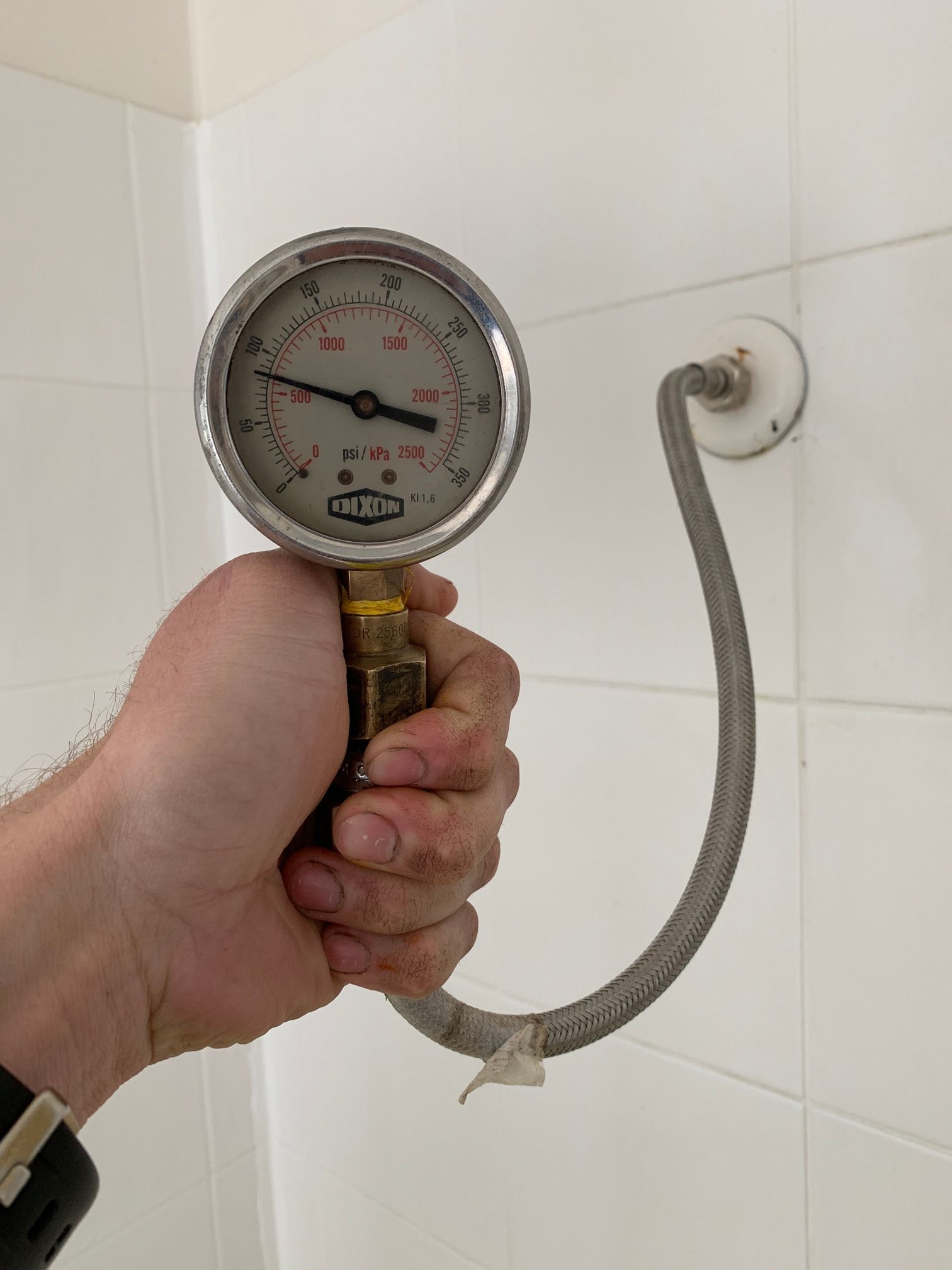 Hand Holding a Pressure Gauge Connected to A Flexible Hose in A Tiled Shower Area — Port Macquarie Plumbing In Port Macquarie, NSW