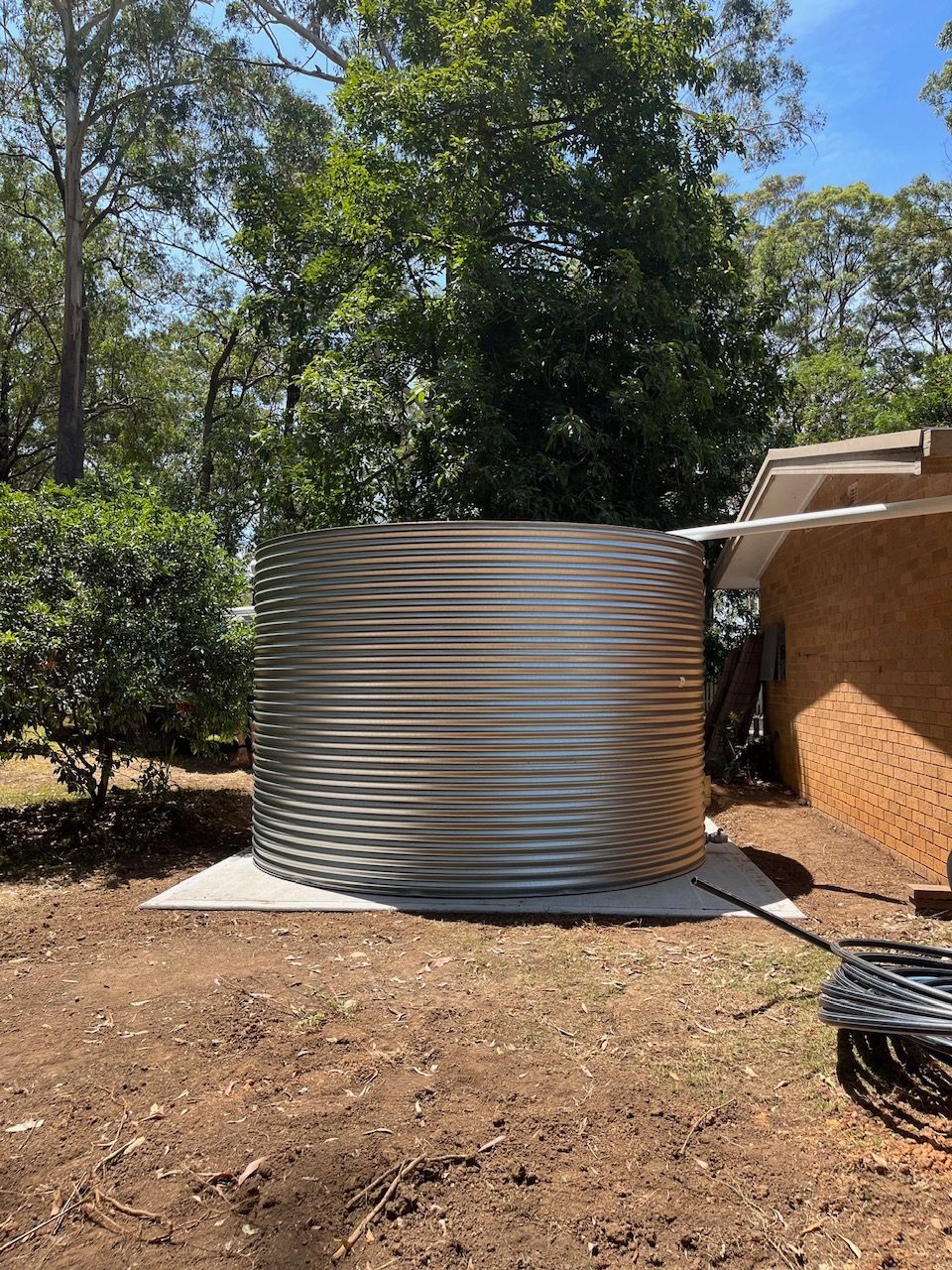 Large Corrugated Metal Water Tank Beside a Building in A Dry Yard with Trees — Port Macquarie Plumbing In Port Macquarie, NSW