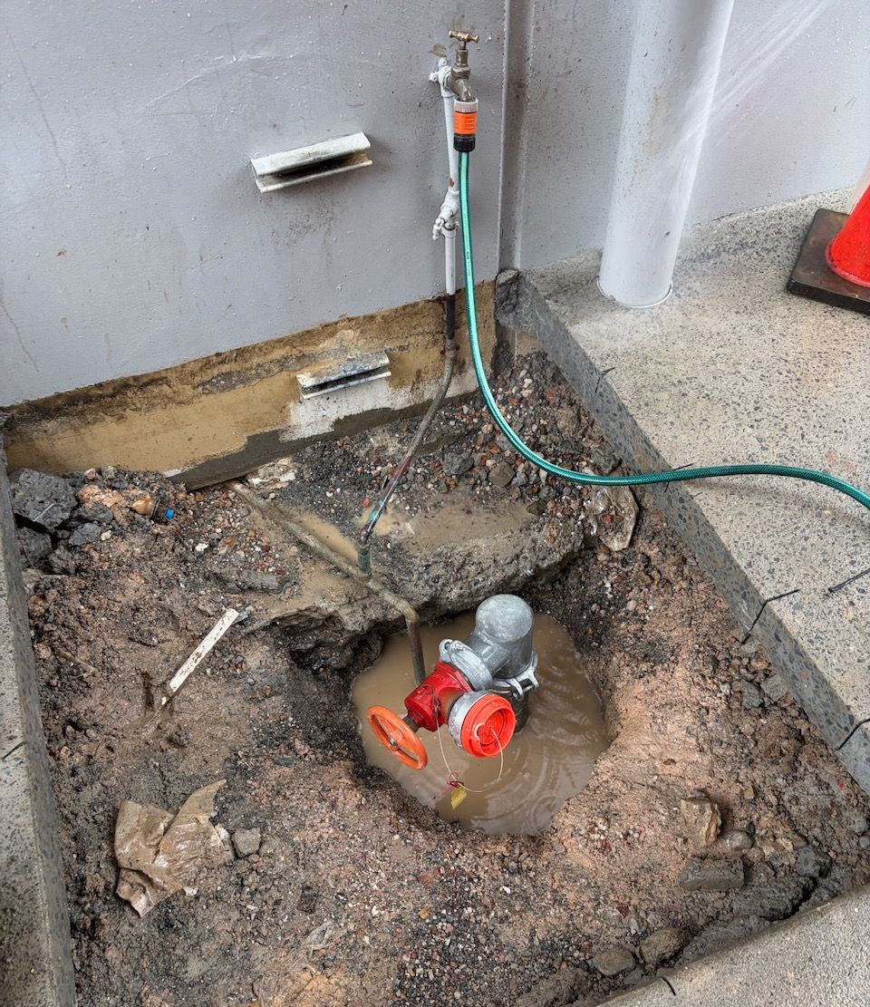 Excavation Pit with Exposed Red Water Valve and Hoses Beside a Wall on Gravel Ground — Port Macquarie Plumbing In Lake Cathie, NSW