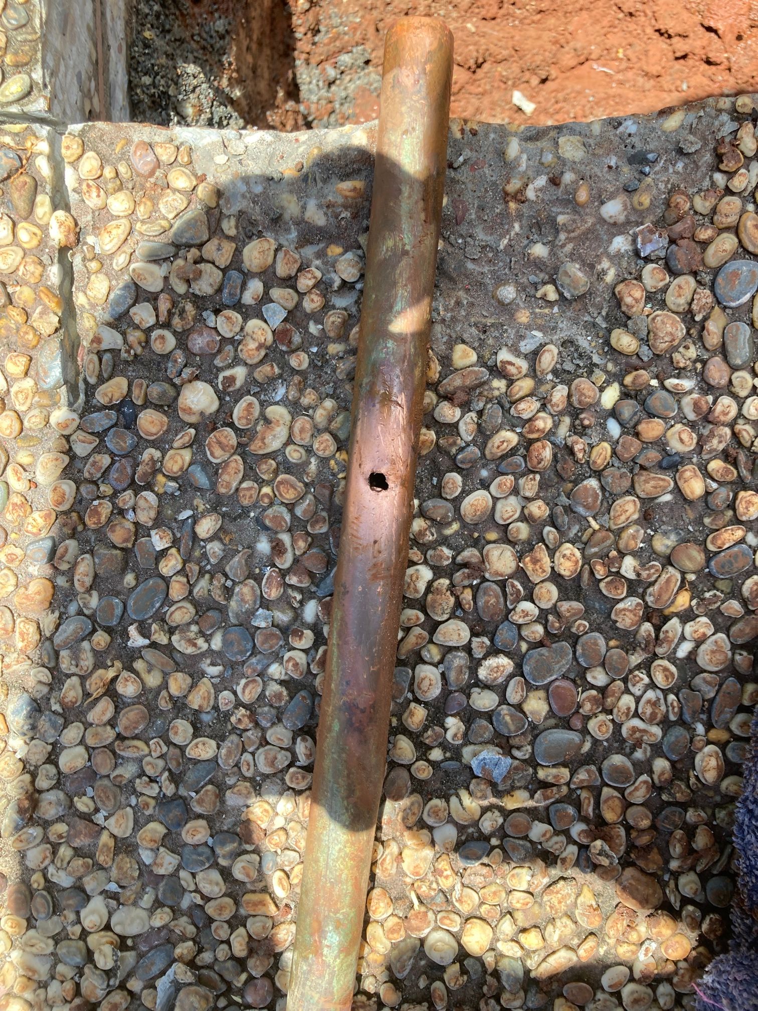A Weathered, Cylindrical Copper Pipe with A Hole Lies on A Bed of Small, Smooth Grey and Brown Pebbles — Port Macquarie Plumbing In Port Macquarie, NSW