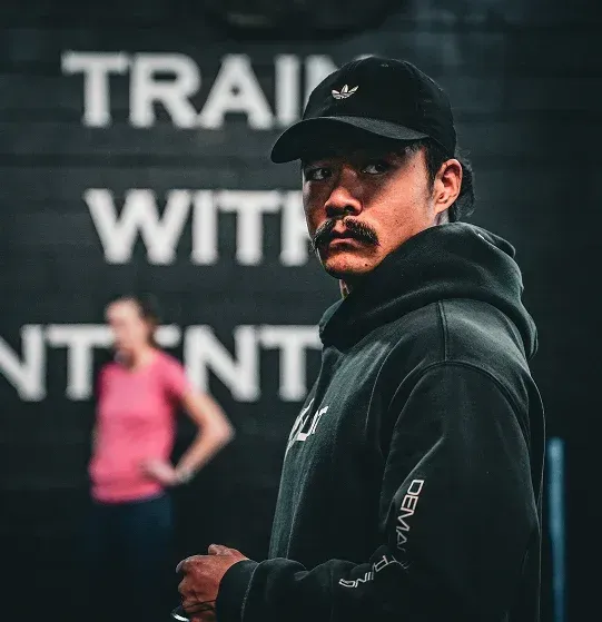 Man in black cap and hoodie looking at camera in a gym, with blurred workout sign and person in pink behind him