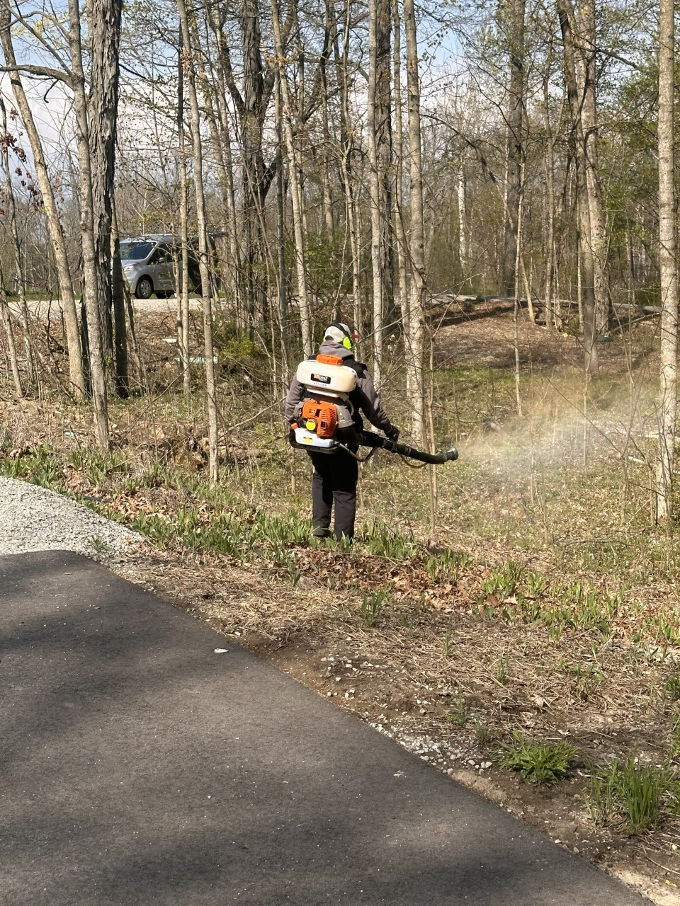 Man using a leaf blower on the edge of a path in a wooded area.
