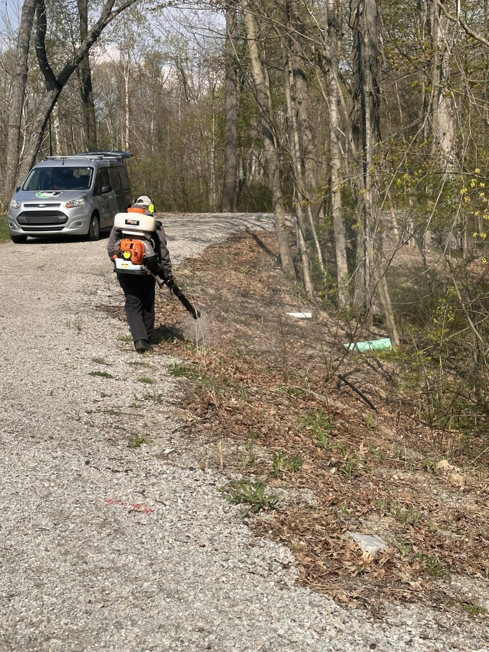 Person using a leaf blower on a gravel path, van parked nearby, trees and foliage on right.