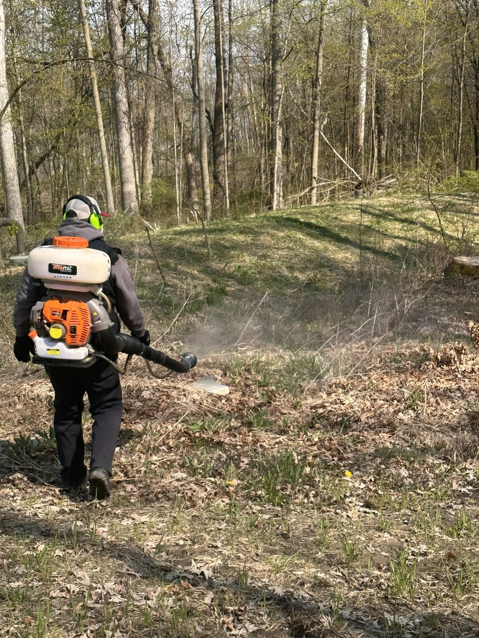 Person using a backpack leaf blower clearing debris in a wooded area.