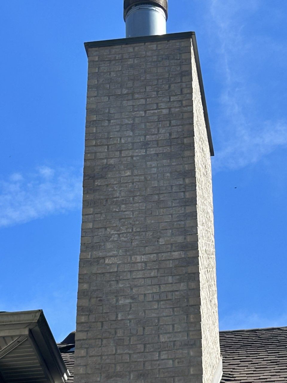 Tall brick chimney against a clear blue sky, with a metal flue and rooftop visible below.