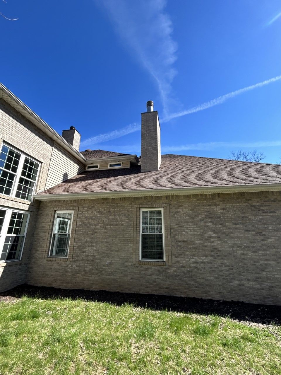 Brick house with brown roof, chimney, windows, and green grass against a blue sky.