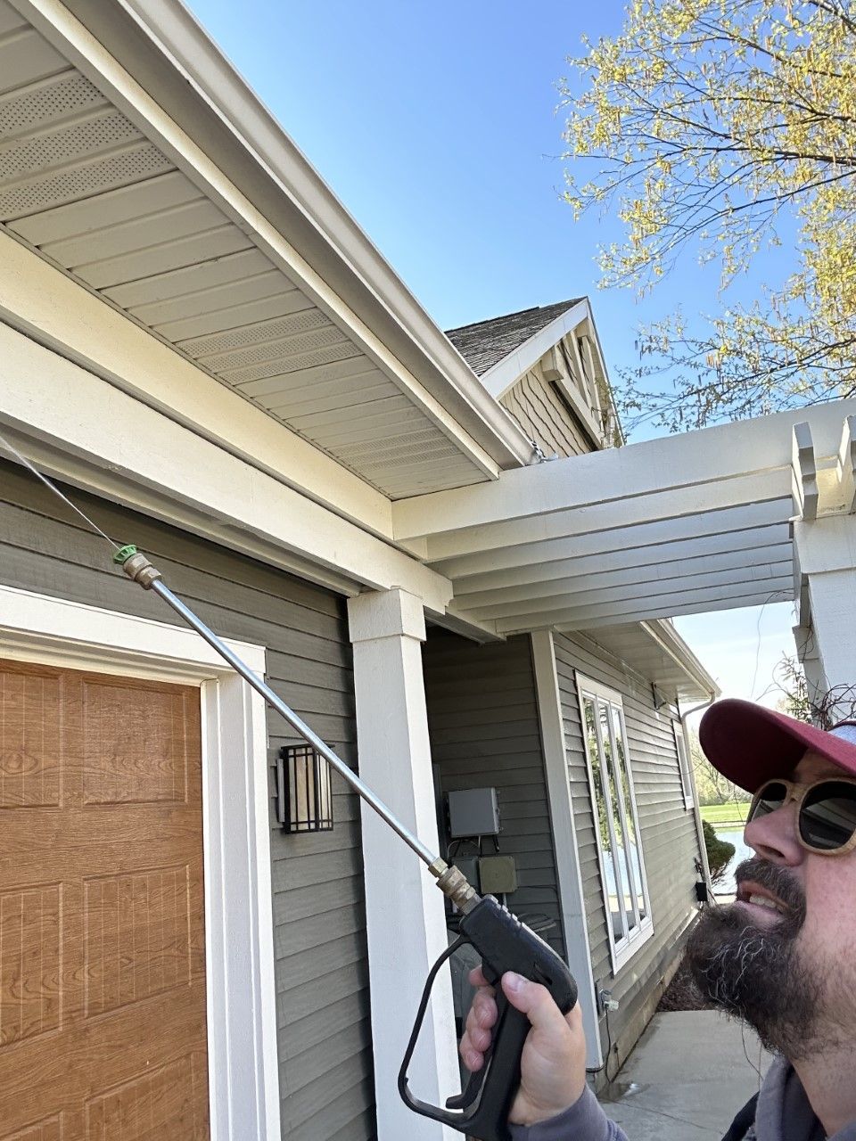 Man power washing siding and soffit of a house with gray siding, white trim, and a brown door.