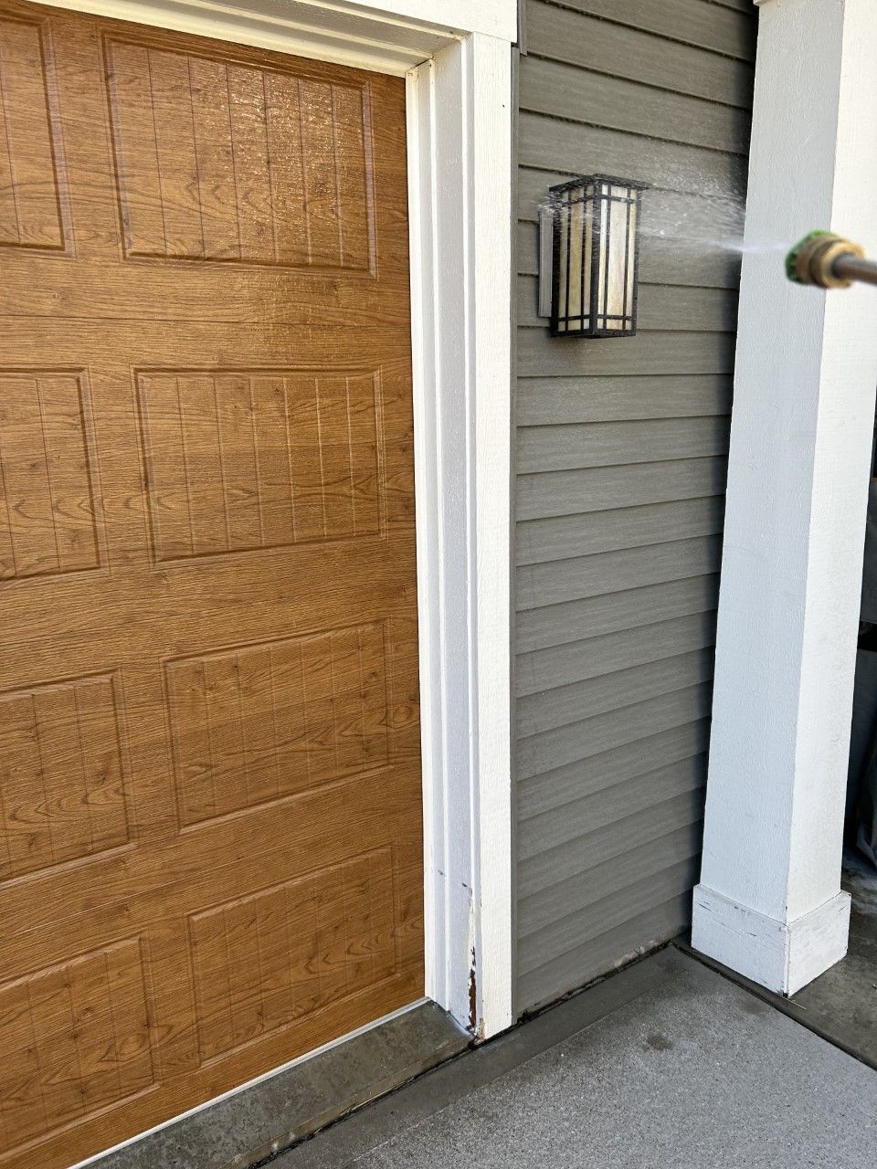 Pressure washing gray siding next to a brown garage door. White trim and a wall-mounted light are visible.
